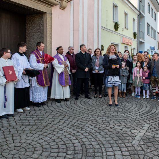 Eine Gruppe von Menschen versammelt sich vor einer Kirche, mit Priestern und einer Frau in einem schwarzen Kleid, die einen roten Stoff und Papier halten. Sie sind von Zuschauern umgeben.