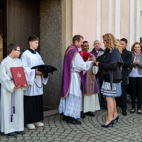 Ein Priester überreicht einer Frau ein Geschenk auf dem Kirchhof, während andere zuschauen.