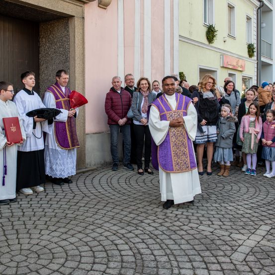 Priester in liturgischen Gewändern stehen vor einer Kirche. Ein Mann in einem violetten Gewand steht vor ihnen und spricht eine Gruppe von Zuschauern an.