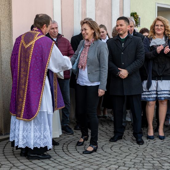 Ein Priester in einer violetten Robe begrüßt eine Frau in einer grauen Blazerjacke, während andere in einem steinernen Bereich vor einem Gebäude zusehen.