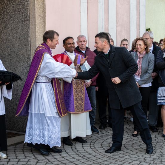 Zwei Priester und ein Mann in einem schwarzen Anzug begrüßen sich mit einem Händedruck vor einer Kirche.