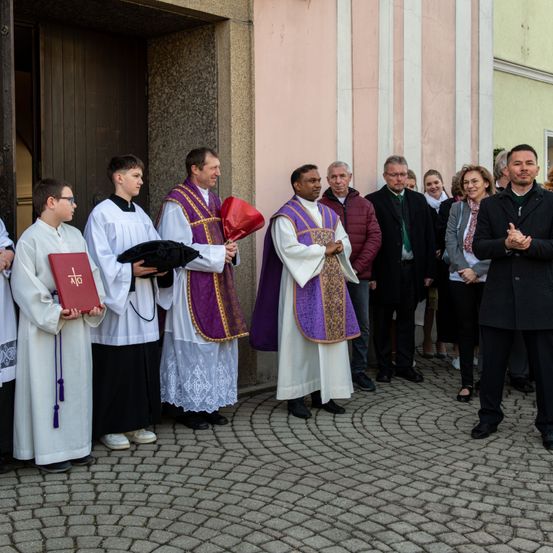 Eine Gruppe von Menschen, darunter Priester und Laien, steht vor einem Kirchengebäude. Die Priester halten Bücher, und einige tragen Ornate. Die Szene deutet auf eine religiöse Veranstaltung hin.