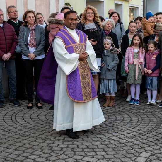 Ein Priester steht lächelnd vor einer Menschenmenge auf einem Kopfsteinpflaster, umgeben von verschiedenen Personen, darunter Kinder und Erwachsene, die alle zu ihm schauen.