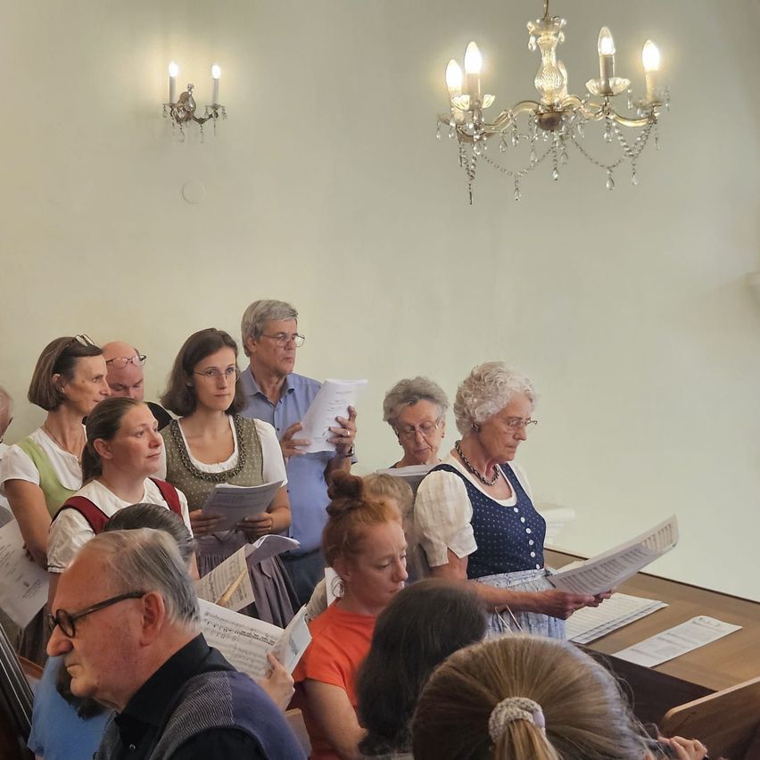 Eine Gruppe von Menschen steht und sitzt in einem Raum und liest von Papieren. Sie scheinen zu singen. Eine Kristallkronleuchter und zwei Wandlampen sind an der Wand befestigt.