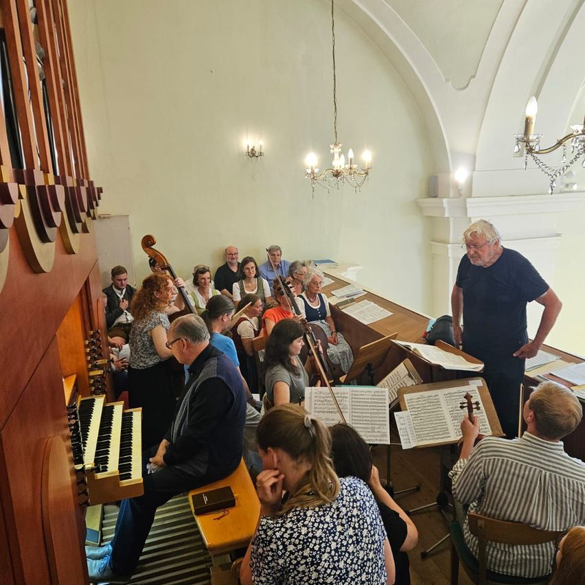 Eine Gruppe von Menschen befindet sich in einem Raum, einige sitzen und andere stehen, um eine Orgel herum. Sie haben Musikinstrumente und Notenblätter. Ein älterer Mann steht vor ihnen.