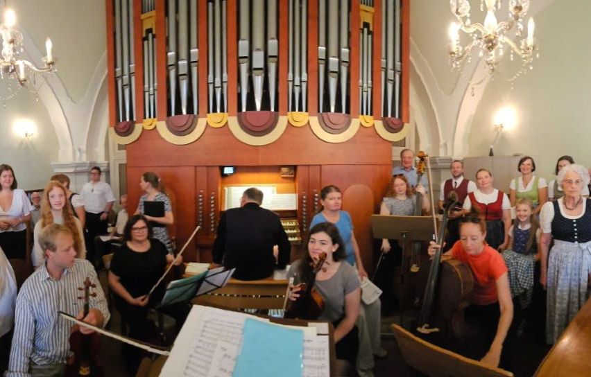 Eine Gruppe von Musikern versammelt sich in einer Kirche, einige spielen Violinen, andere Celli und einer spielt Orgel. Die Notenständer sind mit Musiknoten gefüllt.