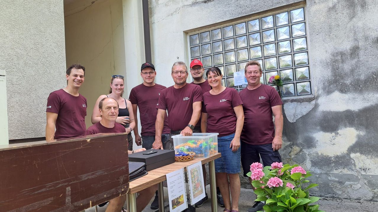 Eine Gruppe von Menschen in weinroten T-Shirts steht vor einem Gebäude. Ein Tisch hält eine Aufbewahrungsbox und Blumen, während sie für die Kamera lächeln.