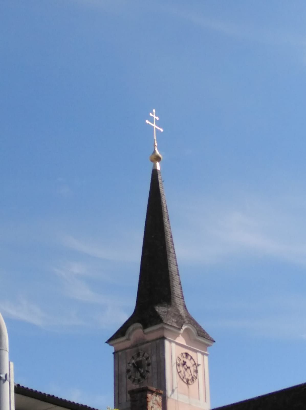 Ein Kirchturm mit einem Kreuz oben und einer Uhr darunter, vor einem blauen Himmel mit Wolken.
