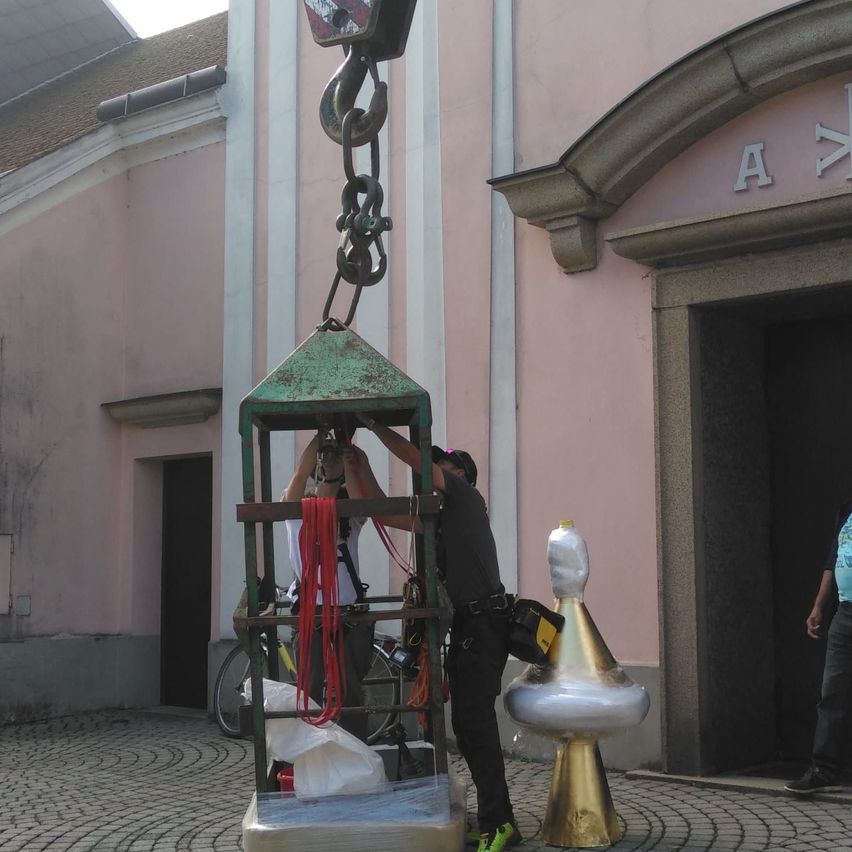 Zwei Personen bereiten sich darauf vor, eine metallische Struktur, wahrscheinlich eine Statue, mit einem Kran vor einem rosa Gebäude mit den Buchstaben AK zu heben.