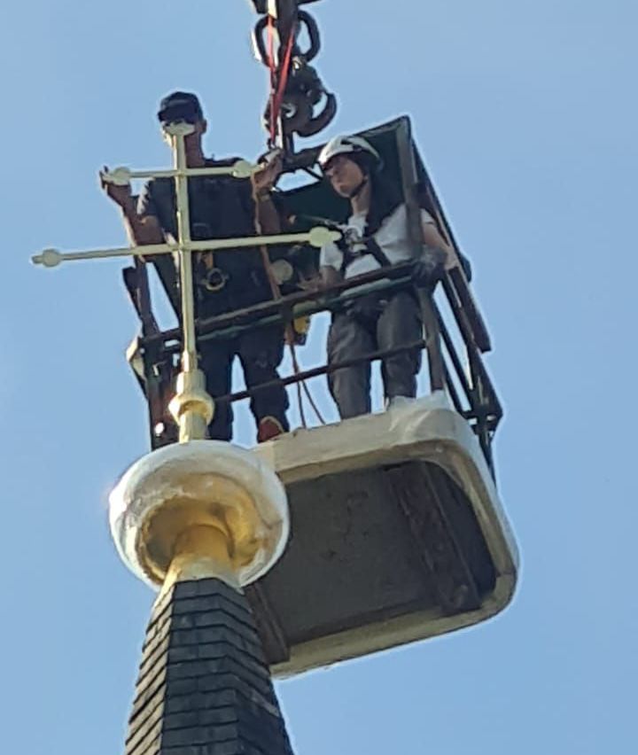 Zwei Personen arbeiten an einer hohen Struktur, eine hält ein Metallkreuz, während die andere auf einer Plattform sitzt, beide tragen Sicherheitsausrüstung und Helme.