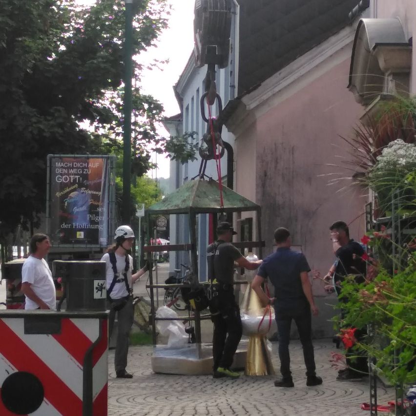 Men versammeln sich um eine Struktur. Ein Mann mit weißem Helm hält eine große Tasche. Ein Gebäude mit einem Banner, das 'MACH DICH AUF DEN WEG ZU GOTT' liest, ist im Hintergrund.