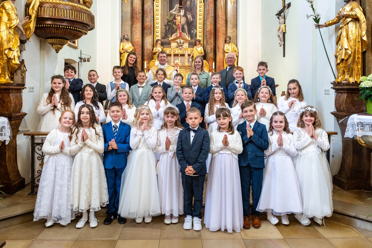 A large group of children dressed in white dresses and suits are standing in a church, praying. There are adults and statues in the background.