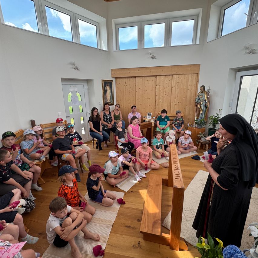 A nun is teaching a large group of children in a church. Some are sitting on the floor, others are sitting on wooden benches. There is a statue of a saint and a picture frame in the background.