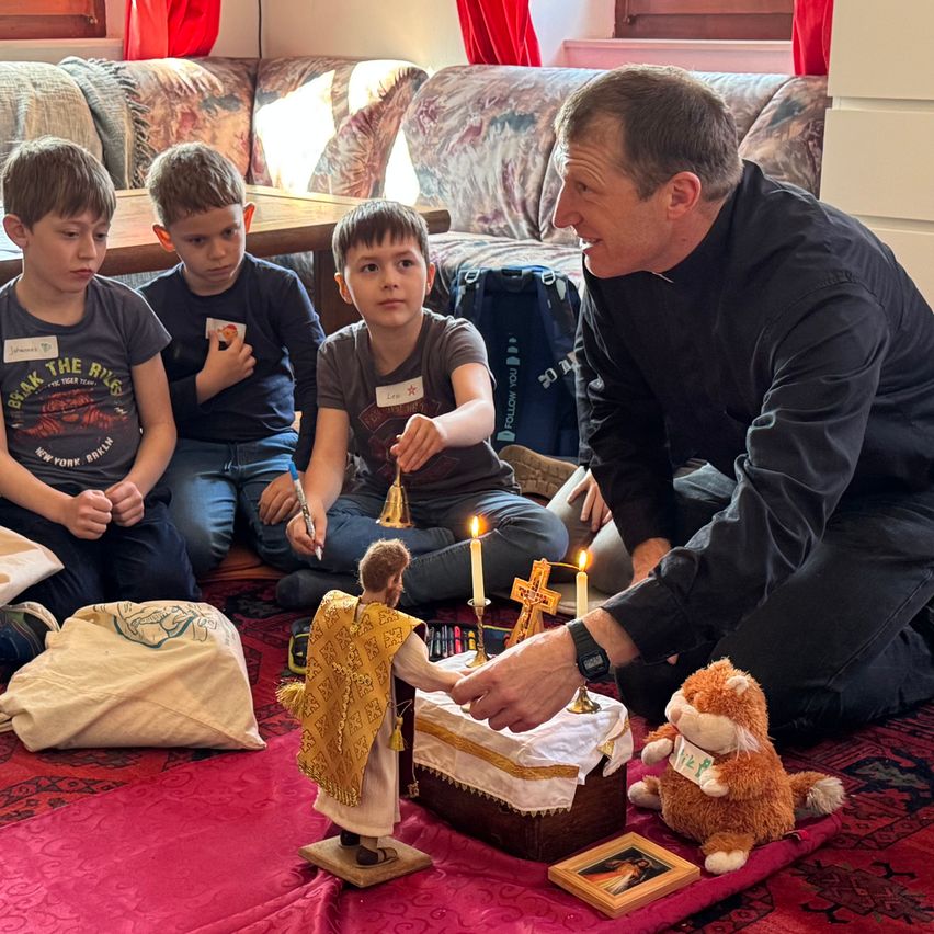 A priest kneels on a carpet, ringing a bell and showing a figurine to three boys, with a cake and candles on a table behind them.