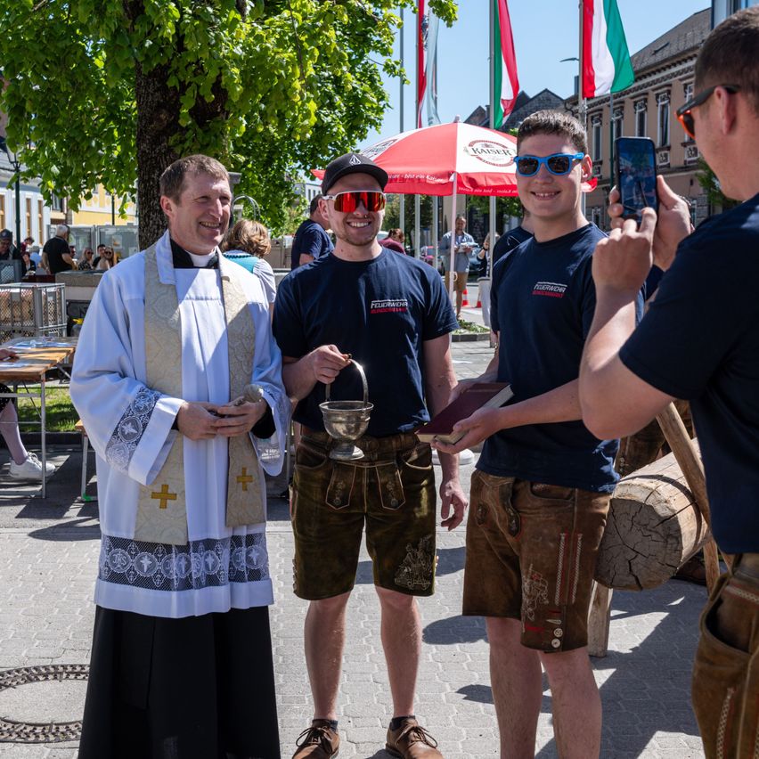 A priest stands with two men, one holding a small cup, while a man takes a photo. Behind them are flags and a tree.