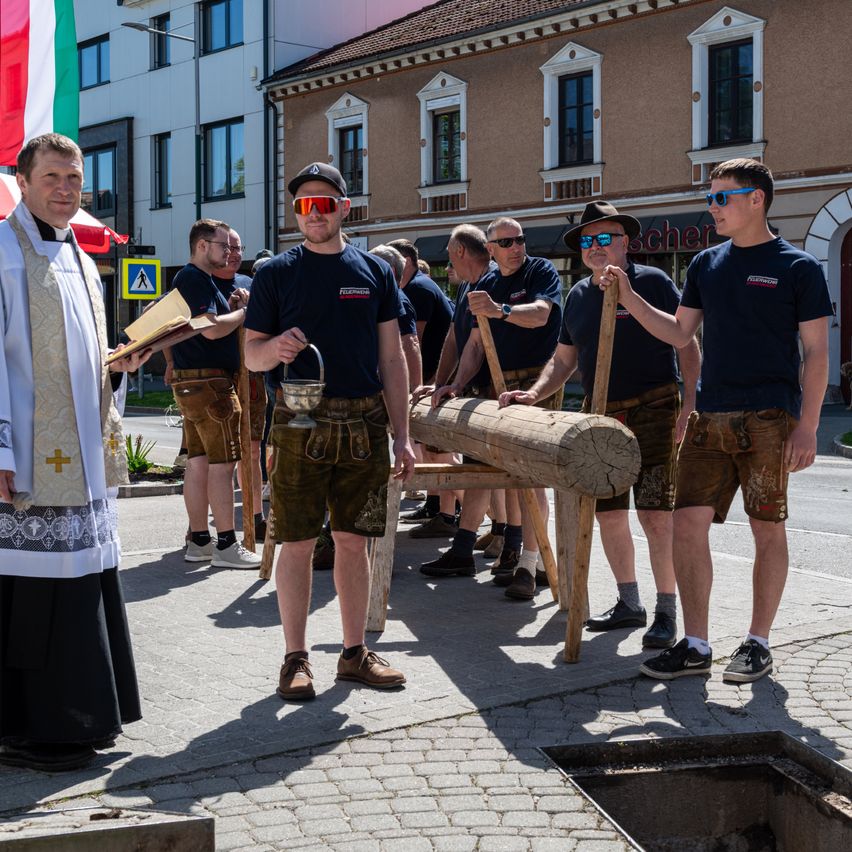 A priest stands beside a group of men in traditional Bavarian attire, holding a log and a lantern, possibly preparing for a ceremonial event.