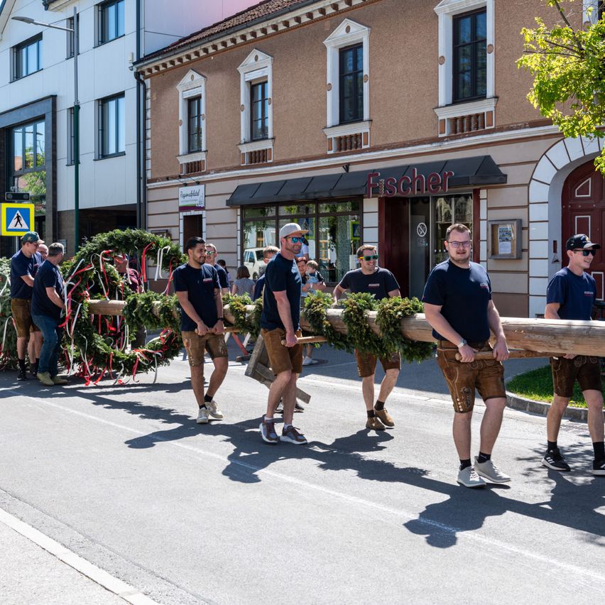 Men in traditional costumes carrying large wooden logs in a parade in front of a building with a sign that reads Fischer.