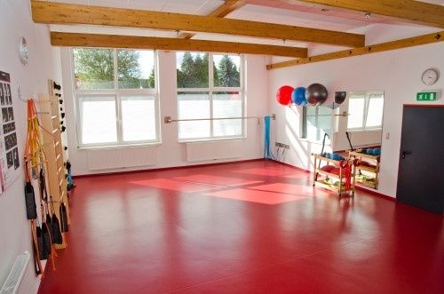 Ein leerer Raum mit rotem Boden und weißen Wänden und Holzbalken. Er hat zwei große Fenster, eine Uhr und einen Spiegel. Es gibt Trainingsgeräte an der Seite.