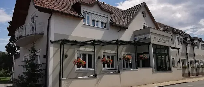 Ein weißes Gebäude mit einem braunen Dach steht an einer Straße mit hängenden Blumentöpfen. Der Himmel ist blau mit weißen Wolken.