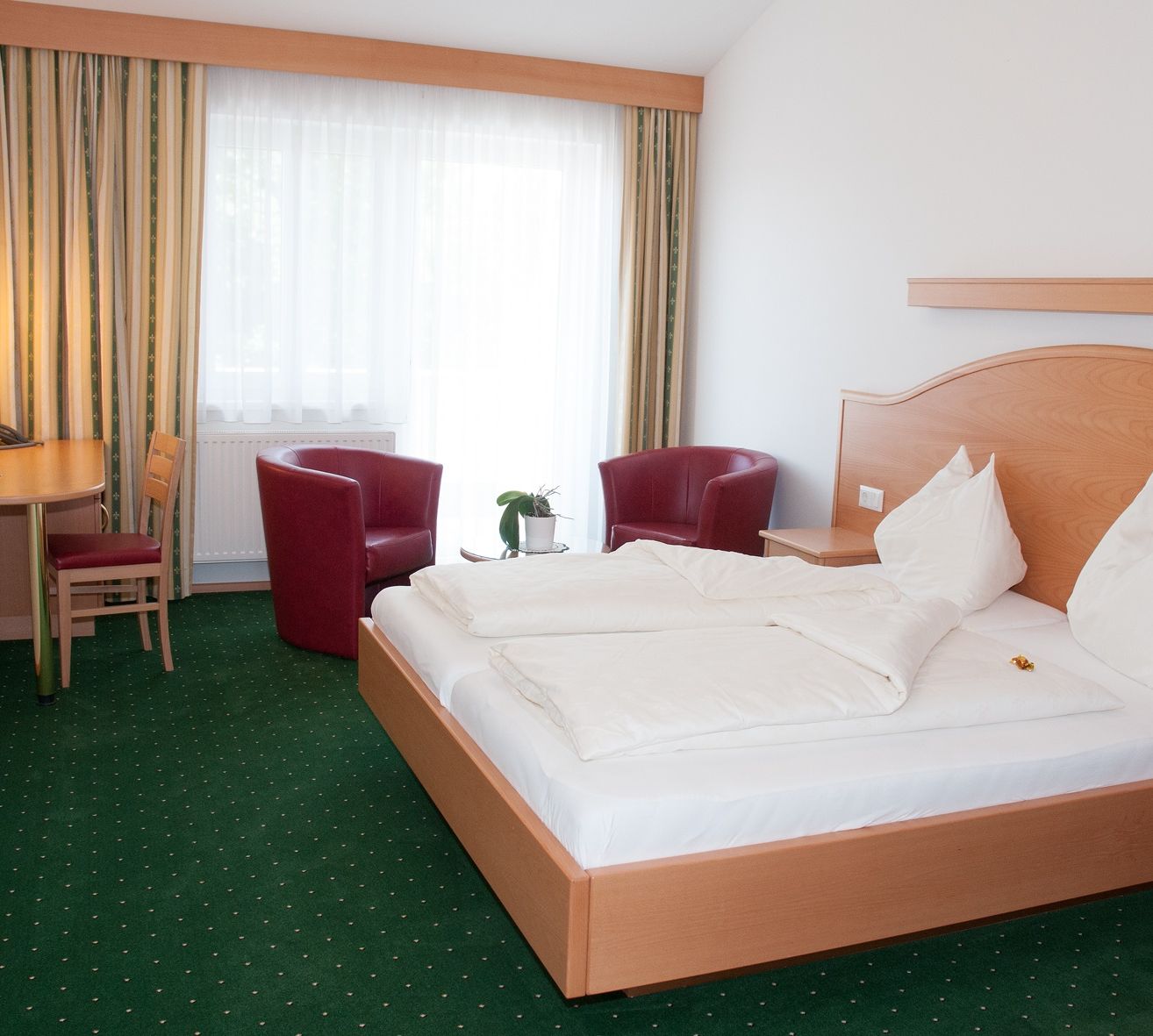 Ein Zimmer mit einem Doppelbett und zwei Stühlen auf beiden Seiten, einem kleinen Tisch in der Ecke und einem Fenster mit Vorhängen.