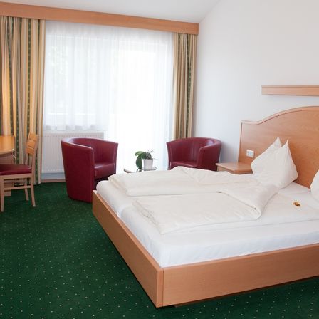 Ein Zimmer mit einem Doppelbett und zwei Stühlen auf beiden Seiten, einem kleinen Tisch in der Ecke und einem Fenster mit Vorhängen.