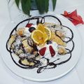 Ein weißer Teller mit einem Dessert aus Bananen, Schlagsahne, Orangenscheiben und Schokoladensirup auf einer weißen Tischdecke. Eine Pflanze steht dahinter.