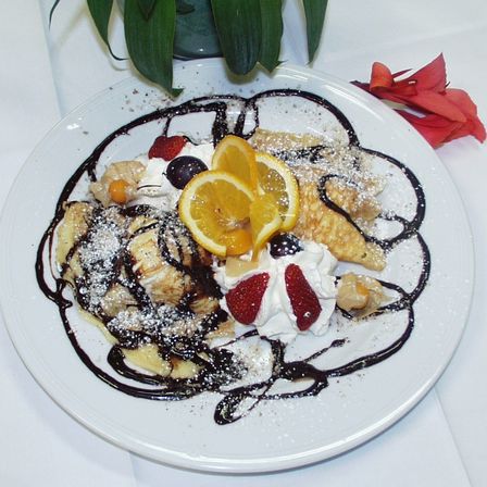 Ein weißer Teller mit einem Dessert aus Bananen, Schlagsahne, Orangenscheiben und Schokoladensirup auf einer weißen Tischdecke. Eine Pflanze steht dahinter.