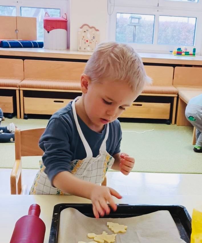 Ein junger Junge in einem blauen Shirt und einer Schürze arbeitet an einem Backprojekt an einem Tisch in einem Klassenzimmer, mit einem Tablett und Backpapier vor ihm.