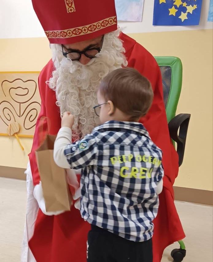 Ein junger Junge mit Brille und einem Hemd mit dem Text Deep Ocean Crew streckt seine Hand aus, um ein Geschenk von einer Person in Santa Claus-Kleidung in einem Klassenzimmer zu erhalten.