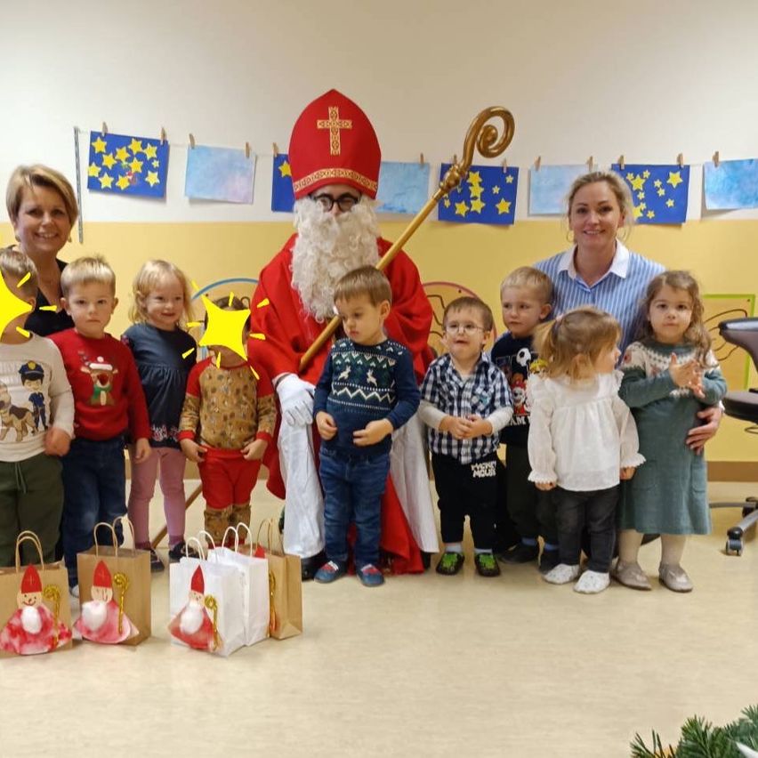 Eine Gruppe von Kindern steht mit dem Weihnachtsmann und zwei Frauen in einem Klassenzimmer mit einer festlichen Atmosphäre, umgeben von Weihnachtsdekorationen und Geschenken.