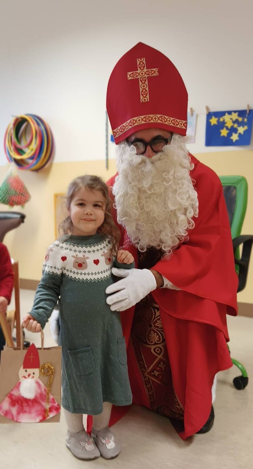 Ein kleines Mädchen steht neben einer Person, die als Weihnachtsmann verkleidet ist, und hält einen Papierbeutel. Die Person trägt einen roten Umhang und einen weißen Bart. Hinter ihnen hat eine Wand eine Flagge und andere Dekorationen.