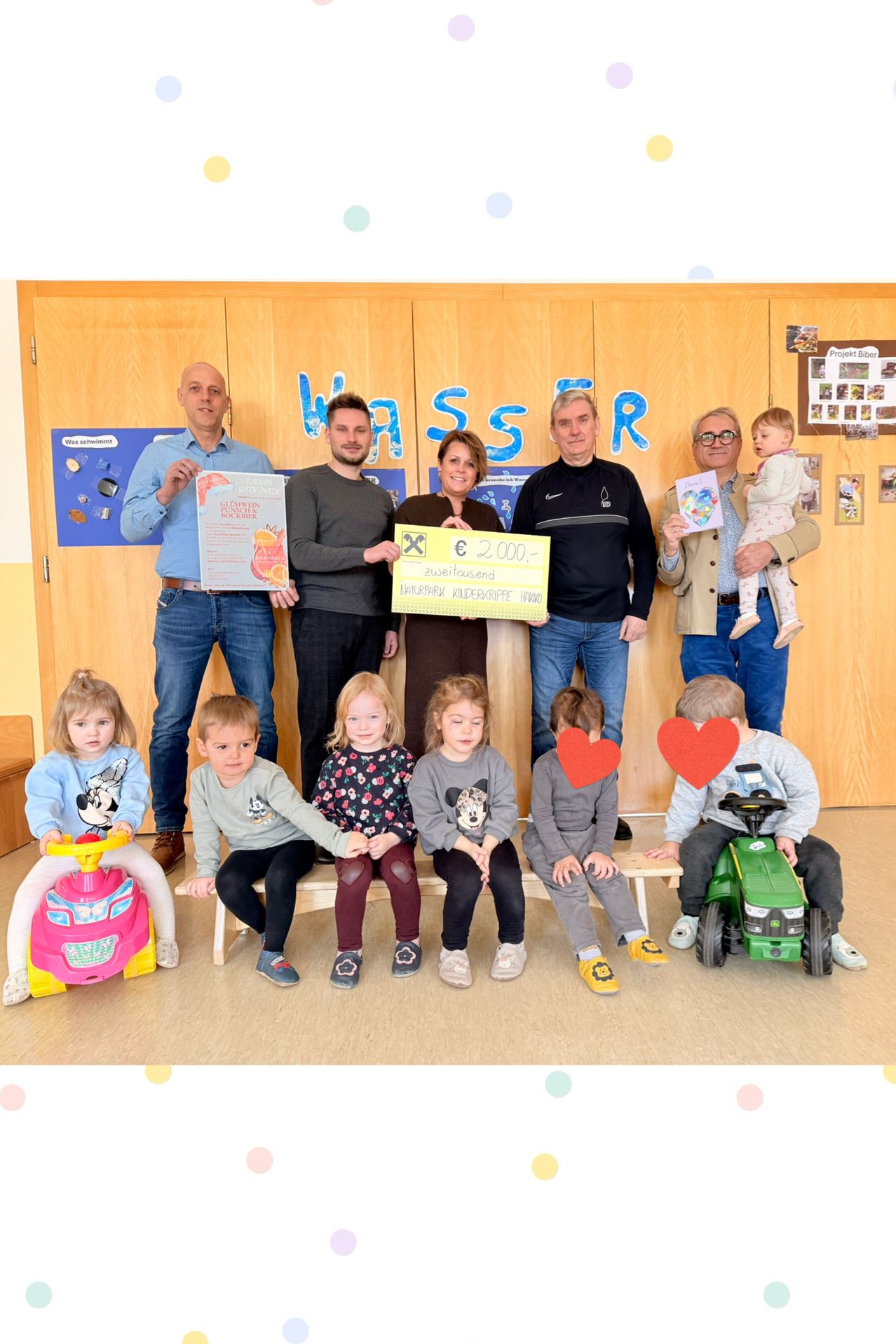 Eine Gruppe von Erwachsenen und Kindern steht vor einer hölzernen Wand mit blauen Buchstaben. Die Erwachsenen halten einen Scheck über 2.000 Euro. Die Kinder sitzen auf Bänken mit Spielzeug.