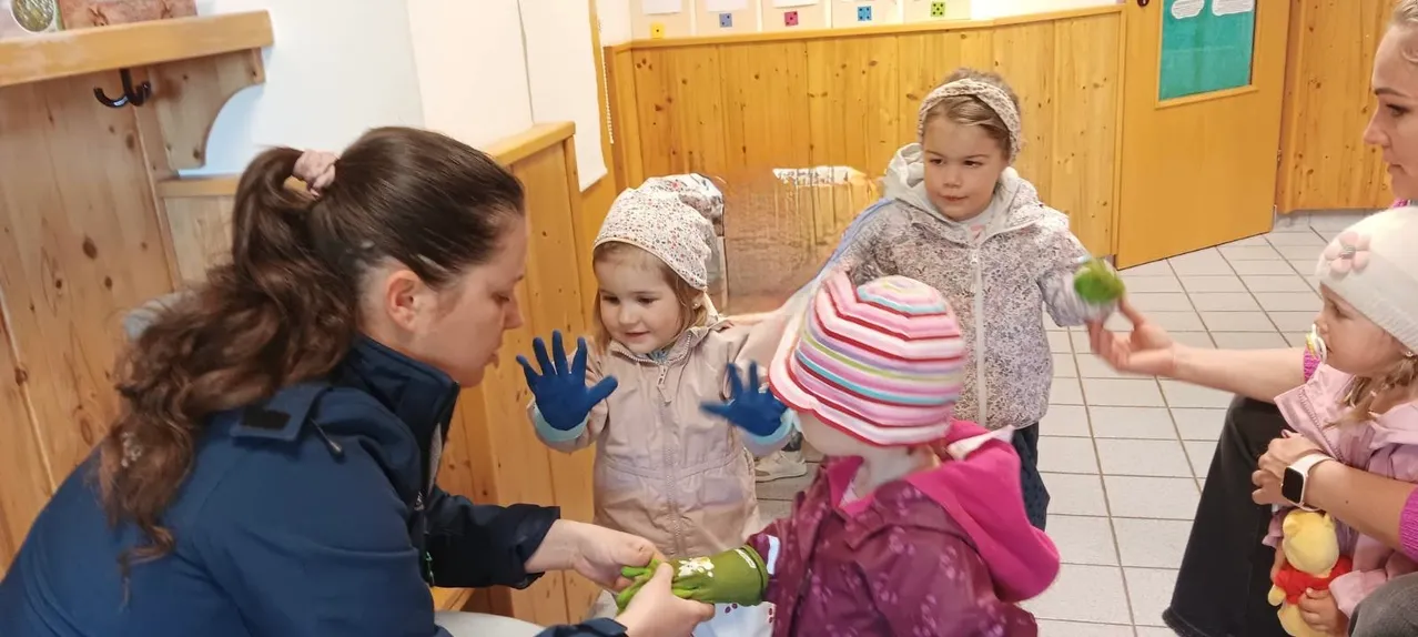 Eine Frau in einer dunkelblauen Jacke hilft zwei kleinen Mädchen mit Handschuhen, die Handschuhe anziehen, in einem Spielzimmer. Die Mädchen tragen Winterjacken und Mützen.