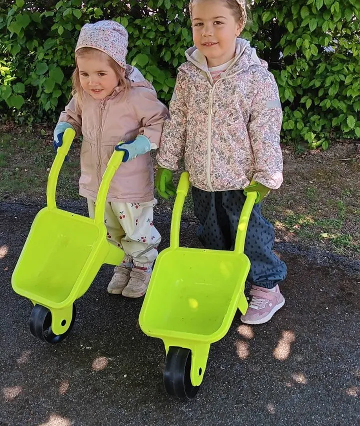 Zwei Kinder stehen auf einem Einfahrtsweg und halten neongrüne Schubkarren. Eines trägt einen rosa Mantel und das andere ein Blumen-Jacke. Sie lächeln beide.
