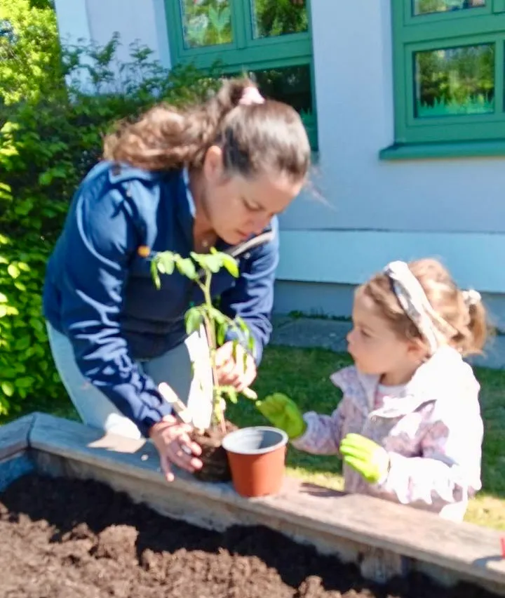 Eine Frau und ein junges Mädchen pflanzen eine kleine Topfpflanze in einem Garten mit einem weißen Gebäude im Hintergrund.