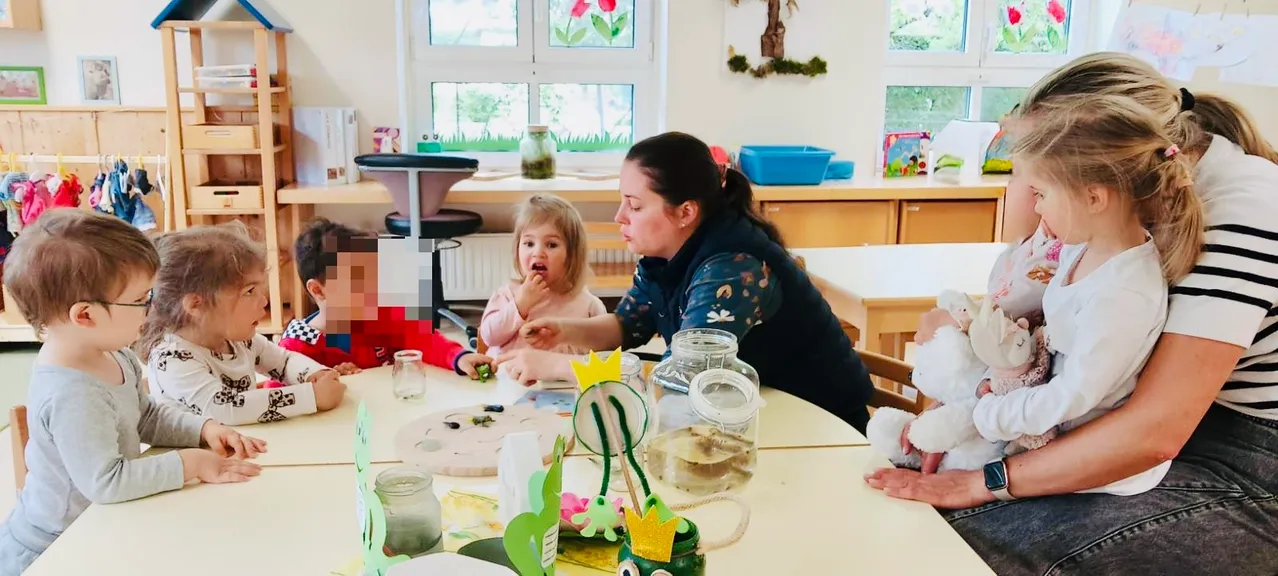 Eine Frau und zwei Kinder sitzen an einem Tisch mit Gläsern, Blumen und Bastelarbeiten. Die Frau beobachtet das Mädchen beim Essen.