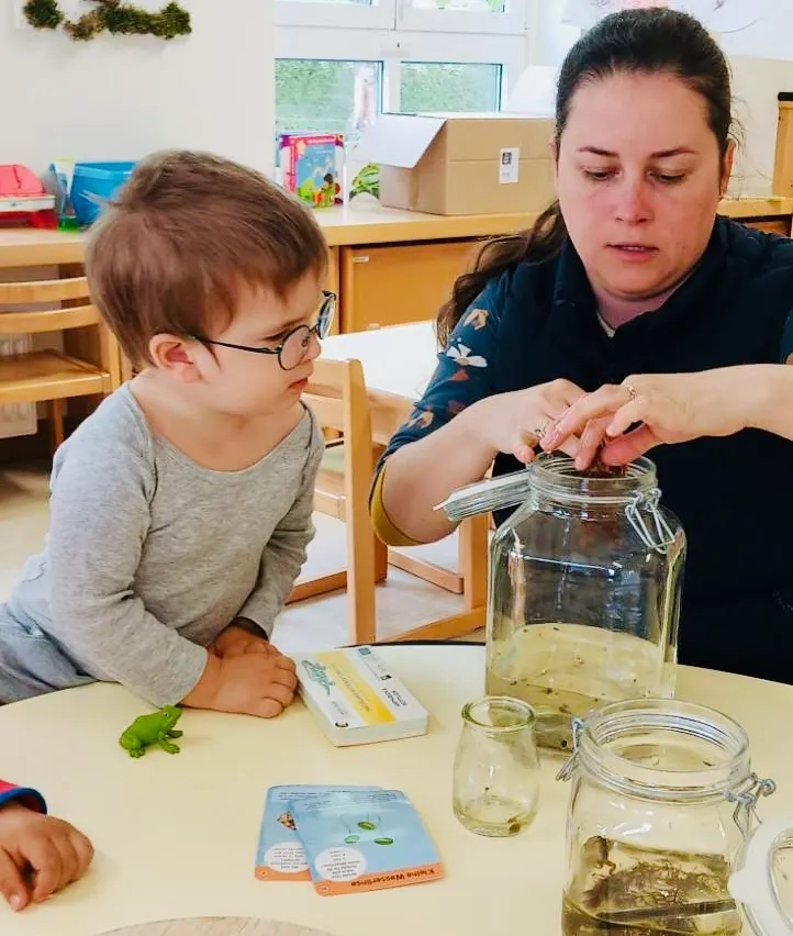 Ein junger Junge mit einer Brille beobachtet, wie eine Frau einen Frosch in ein Glas setzt. Ein Tisch hat Gläser und Bücher. Sie sind in einem Raum mit Holzmöbeln.