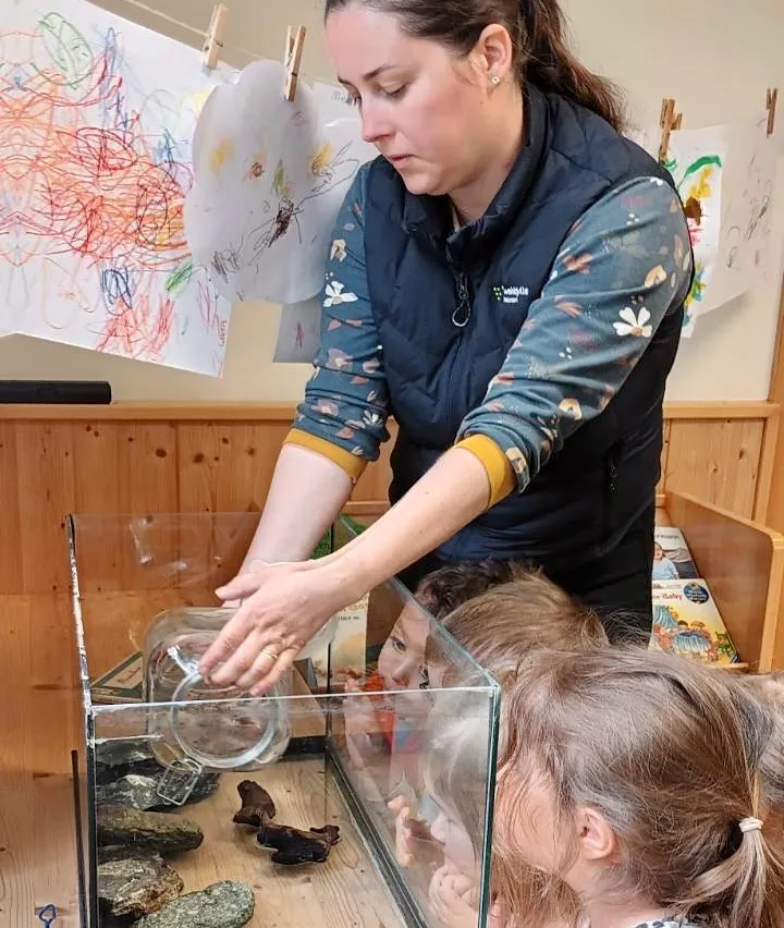 Eine Frau hält einen Glasbehälter mit Wasser vor zwei Kindern. Sie erklärt etwas im Inneren des Behälters. Die Kinder schauen in den Behälter.