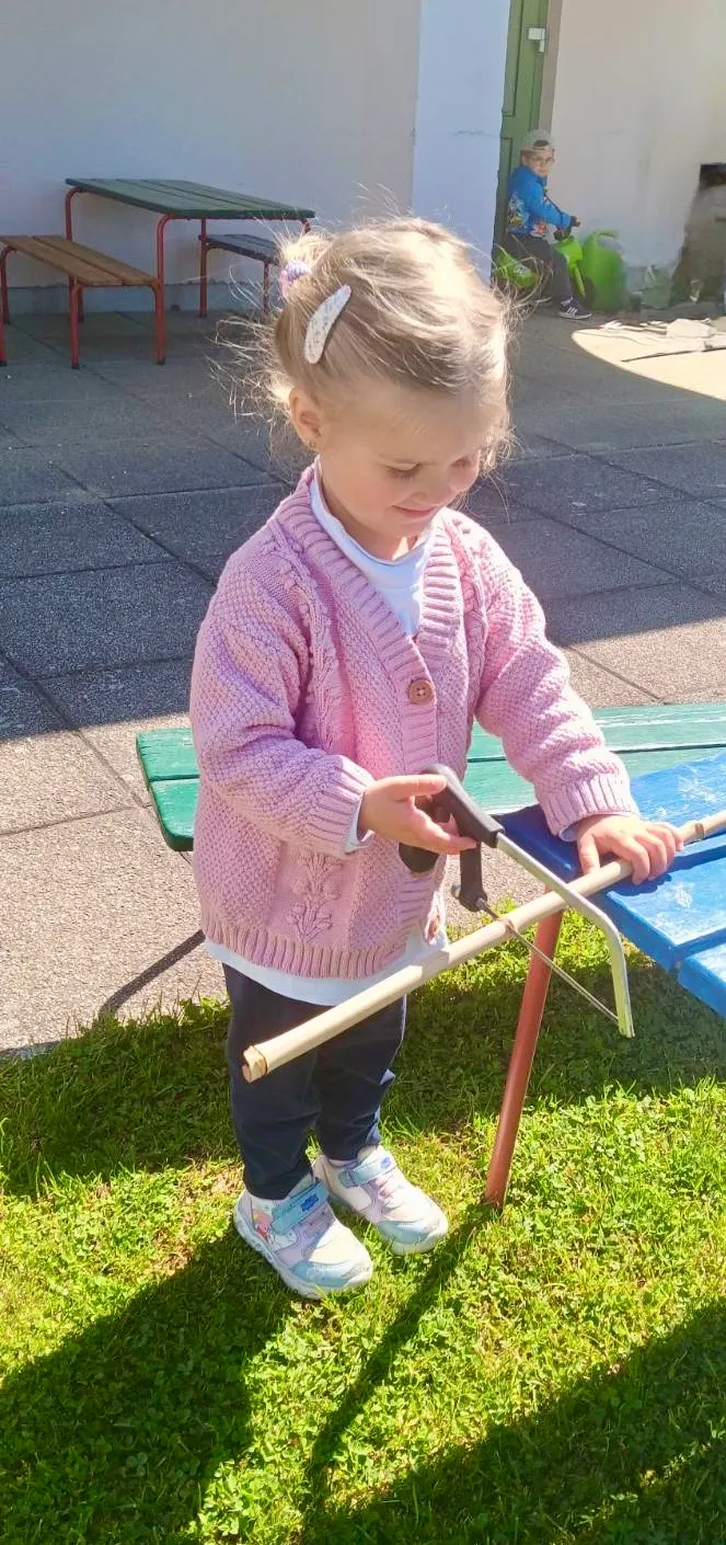Ein kleines Mädchen in einem rosa Pullover steht auf Gras und schneidet mit einer Säge einen Holzstab. Sie konzentriert sich auf ihre Aufgabe.