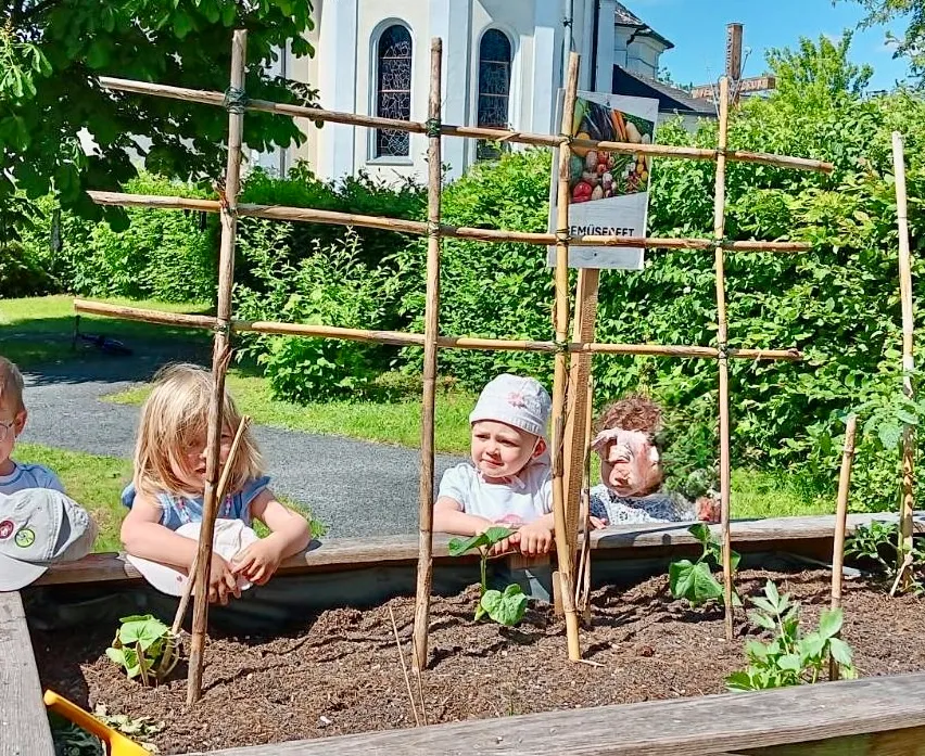 Drei Kinder schauen durch einen Bambusrahmen in einem Garten. Ein Schild mit Gemüse ist am Rahmen befestigt. Ein Gebäude und üppiges Grün im Hintergrund.
