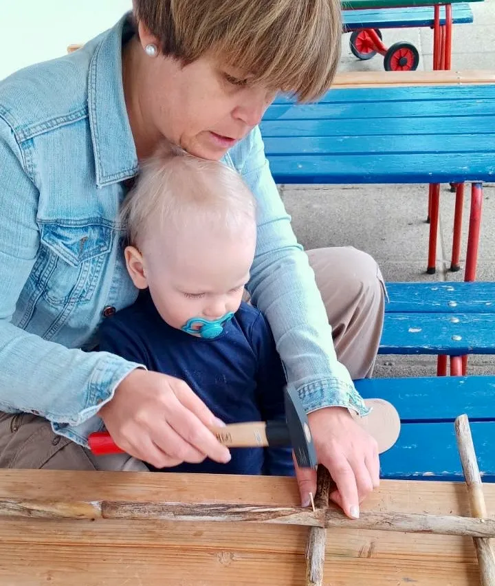 Eine Frau und ein Kleinkind sind zusammen, wobei die Frau einen Hammer auf einer Holzplanke benutzt, während das Kind aufmerksam zusieht. Der Ort scheint sich im Freien zu befinden.