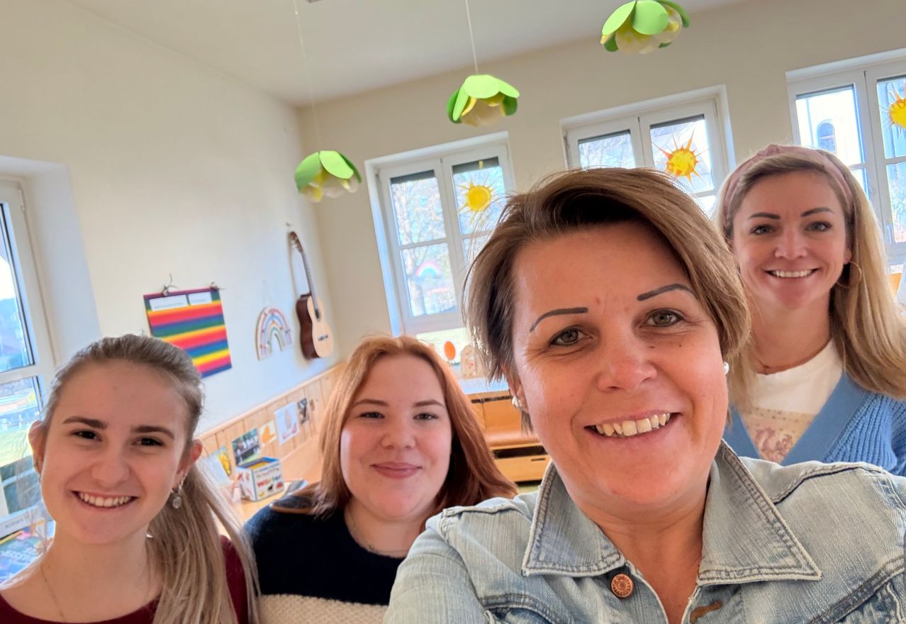 Vier Frauen lächeln in einem Raum mit dekorativen hängenden Blumen, einer Gitarre und einem Regenbogen. Die Frau vorne trägt eine Jeansjacke.