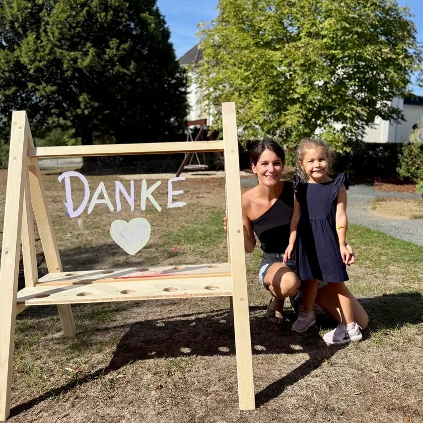 Eine Frau und ein junges Mädchen stehen neben einem Holzschild mit dem Wort Danke darauf. Das Schild hat ein Herz darauf gezeichnet. Sie lächeln und posieren für ein Foto.