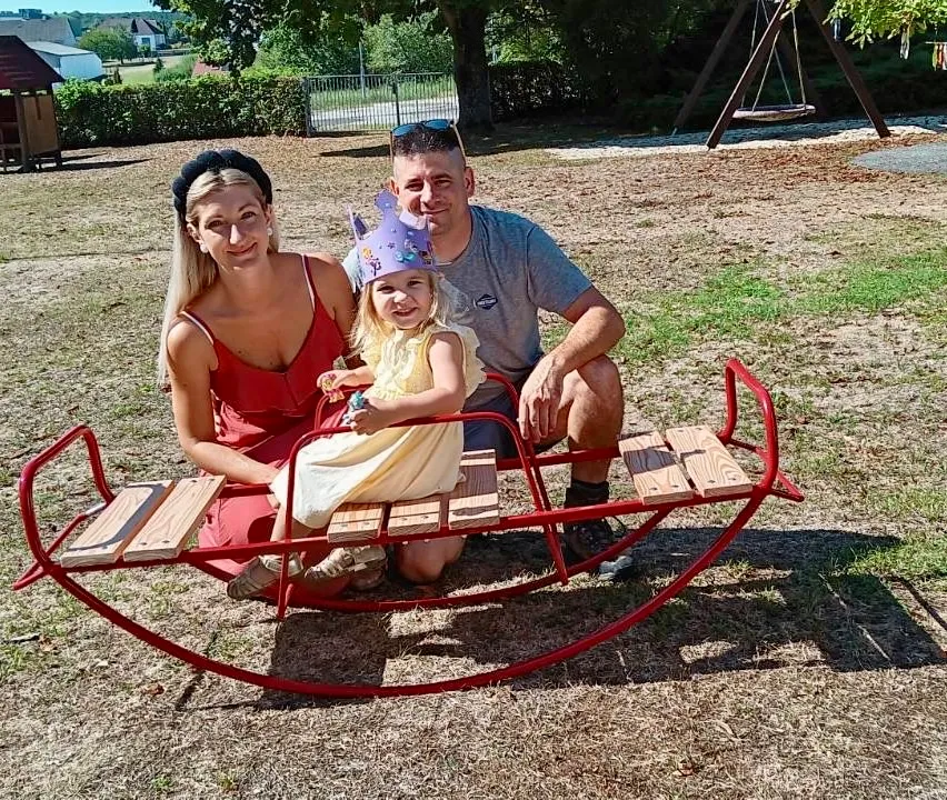 Eine Familie aus drei Personen, eine Frau, ein Mann und ein junges Mädchen, sitzen auf einem roten Schlitten in einem Park. Das Mädchen trägt eine lila Krone.