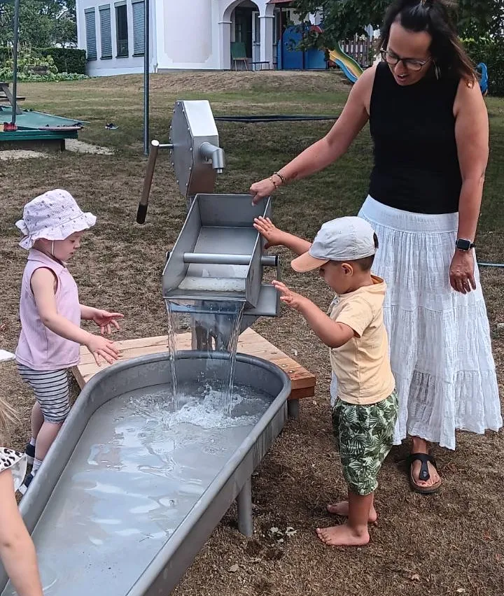 Eine Frau und zwei Kinder spielen mit einer Wasserfontäne in einem Park. Die Kinder sind barfuß und die Frau trägt ein schwarzes Tanktop und eine weiße Rock.