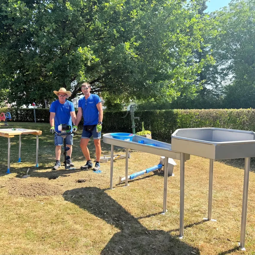 Zwei Männer in blauen Hemden und Shorts stehen in einem Garten, einer hält ein Werkzeug und der andere trägt einen Hut. Sie sind von Gartenwerkzeugen und einem kleinen Tisch umgeben. Im Hintergrund befinden sich Bäume und eine Hecke.