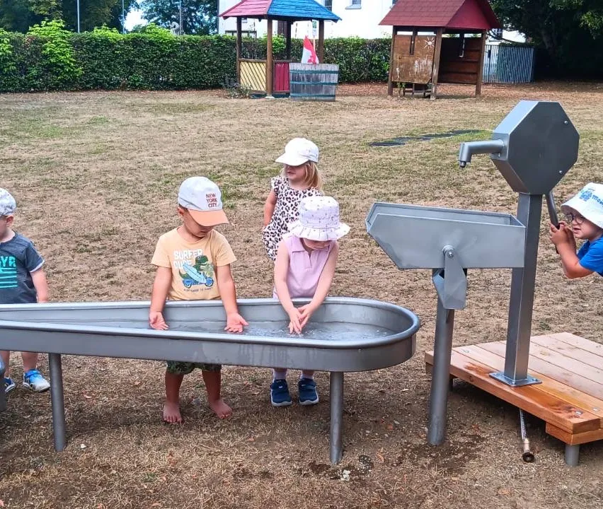 Drei Kinder spielen mit Wasser in einer großen Metallwanne, mit einer Wasserpumpe in der Nähe. Dahinter befindet sich ein Spielplatz mit kleinen Häusern.