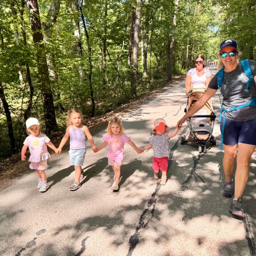 A woman with a backpack leads a group of young girls holding hands on a wooded path. Behind them, a woman pushes a stroller.
