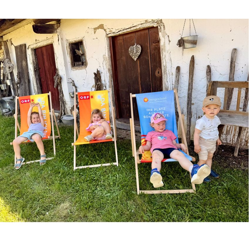 Four kids are sitting on chairs in front of a building with a blue banner on one of the chairs.