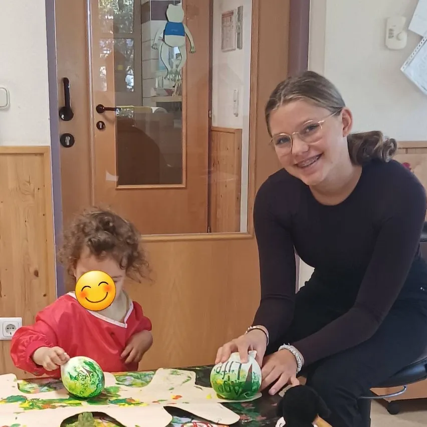 Eine Frau und ein Kind malen Kürbisse auf einem Tisch. Das Kind hat ein lächelndes Gesicht auf der Wange.